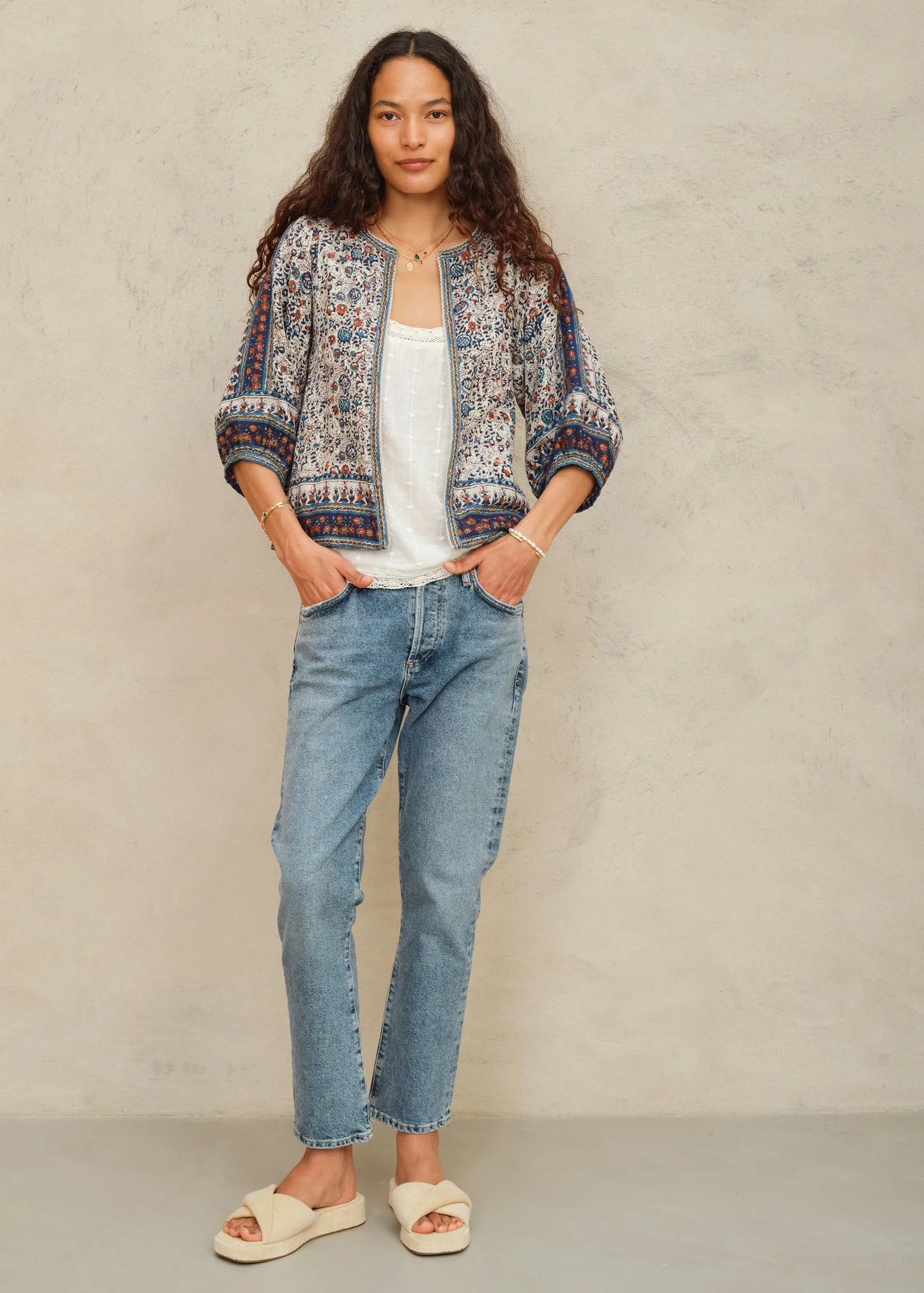 Woman wearing a patterned jacket, white top, and blue jeans against a beige background