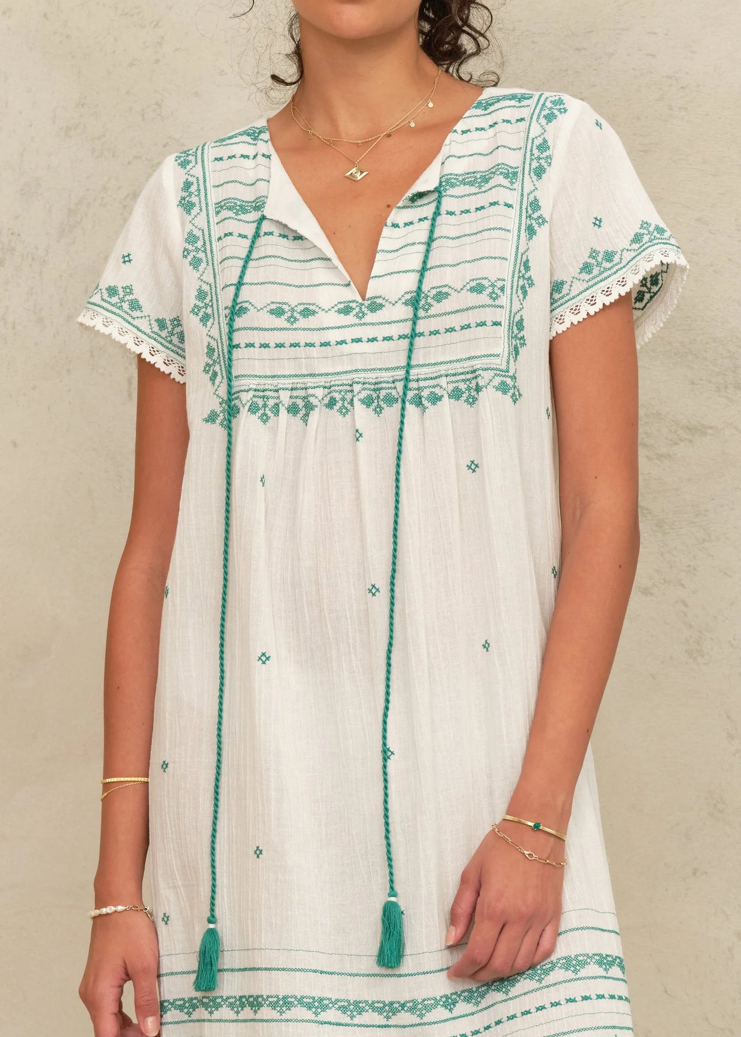 Woman wearing a white dress with green embroidery and tassels against a neutral background