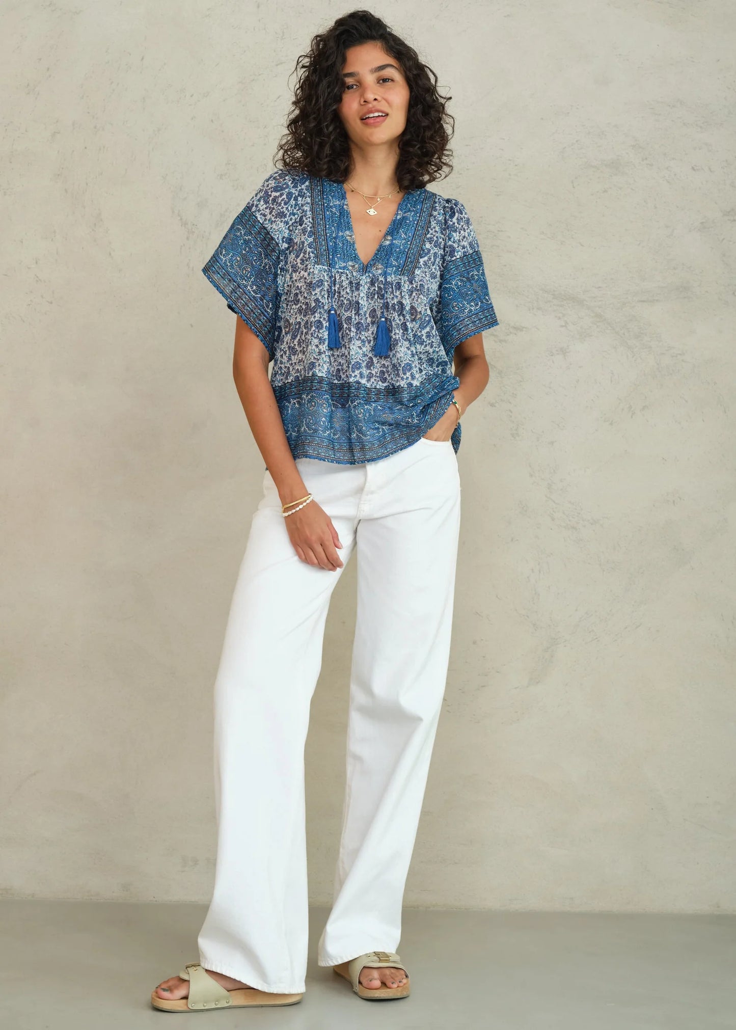Woman wearing a blue patterned top and white pants against a beige background