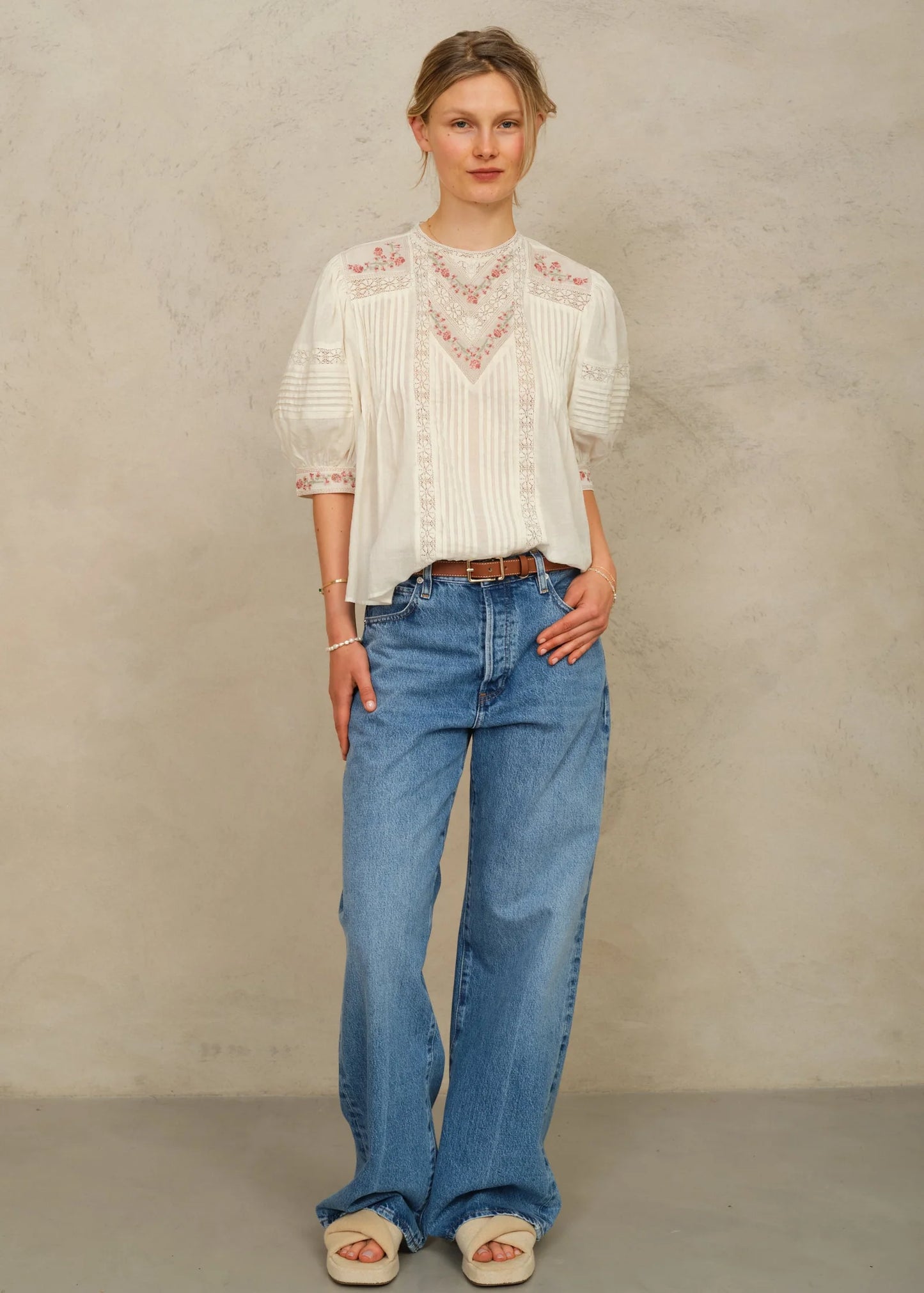 Woman wearing a white blouse with lace details and blue jeans against a beige background