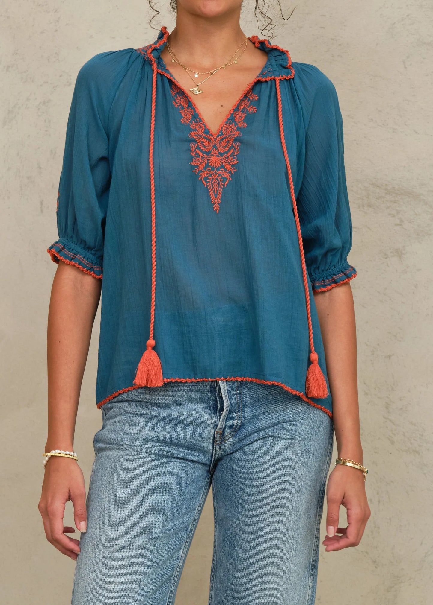 Woman wearing a blue blouse with orange embroidery and tassels, paired with light blue jeans against a neutral background.